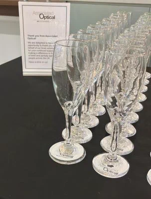 2 rows of wine glasses resting on discs with Associated Optical partner of Eschenbach. On the left side is a note from Associated Optical thanking everyone for supporting blind and partially sighted people, and offering a drink on them. 