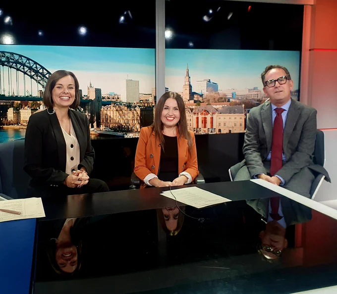 Two women and a man are sat at a newsdesk smiling to camera. Behind them is an image of the Tyne Bridge.