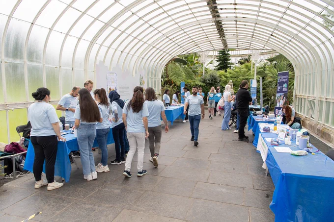 A series of activity tables with blue table cloths lined up in the Kibble palace in the Glasgow Botanic Gardens. There are about 20 people gathering round looking at what is on some of the tables. 