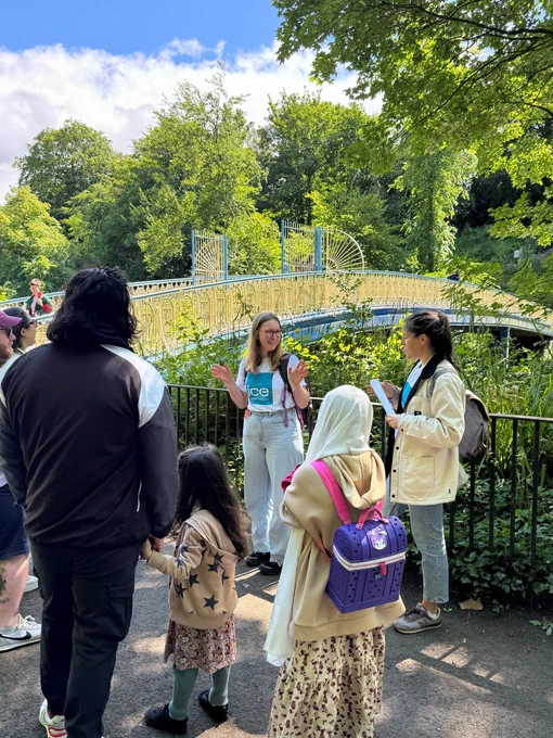 Two people wearing ICE branded T-shirts standing in front of a yellow and blue iron bridge on the River Kelvin with a small group of people watching. 
