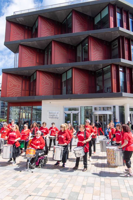 A marching band of 20 people wearing red tshirts and carying drums of various sizes standing outside the Byres Hub building which is 4 stories, made of red material with windows set on 45 degree angles. 