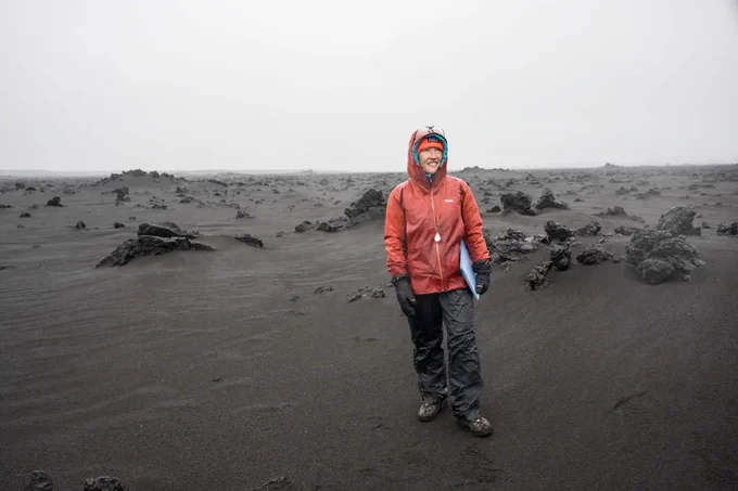 NASA astronaut and Artemis II mission specialist Christina Koch stands in the desolate landscape of Iceland during a geology field training course. Koch is wearing a rust-colored windbreaker and long black pants, both of which are drenched in water. Credit: NASA/Robert Markowitz