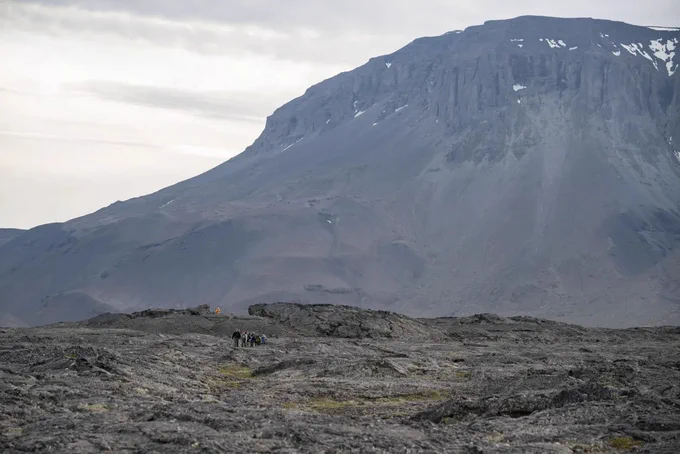 The Artemis II crew and backup crew members trek across the Icelandic landscape during their field geology training. The dark, rocky landscape dominates the image, with the crew barely visible as a group of small figures left of center. Credit: NASA/Robert Markowitz