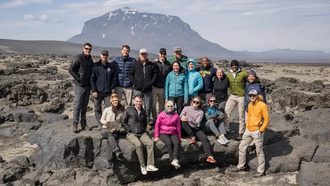The Artemis II crew astronauts, their backups, and the geology training field team pose in a valley in Iceland’s Vatnajökull national park. The group of about twenty is arranged in two loose rows, one standing in the back and one sitting on a large rock in front. The background is grey and rocky, with a large mountain in the distance. Credit: NASA/Robert Markowitz