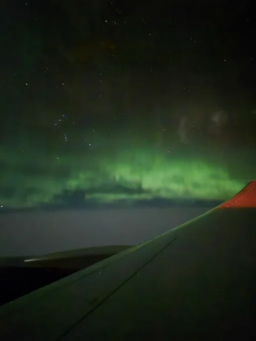 A photograph taken through and aeroplane window showing the wing in the foreground, and a night sky. Several stars are visible, including the constellation of Orion. A bright green glow extends across the sky.