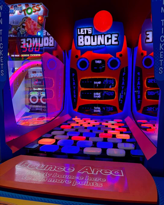 View of an arcade game area featuring a 'Let's Bounce' ball-toss game. The game is lit with vibrant blue and orange lights, displaying scoreboards and numerous colored circles on the playing surface ready for bouncing balls.