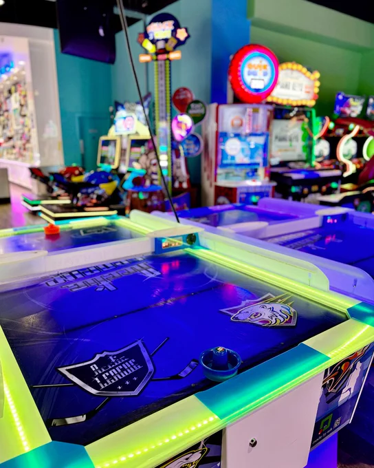 An air hockey table lit with neon green lights, featuring the Buffalo Sabres logo, set in a colorful arcade room with various games and prizes visible in the background.