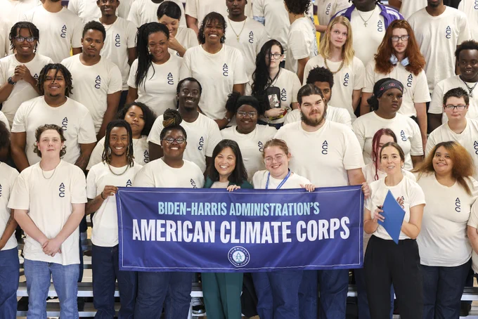 Acting Secretary Julie Su stands in a crowd of smiling young people wearing white shirts. They hold a banner that reads, "Biden-Harris administration's American Climate Corps."