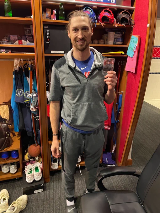 Matt Strahm smiles and poses in front of his locker with an autographed baseball card. He's wearing a gray sweatsuit over a Phillies t-shirt.