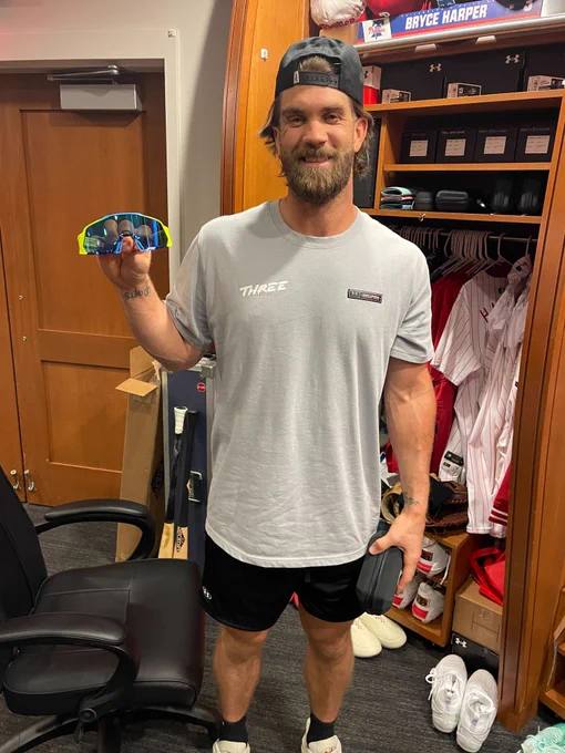 Bryce Harper smiles and poses for a picture in front of his locker while holding a pair of yellow and blue sunglasses. He's wearing a backwards hat, a gray t-shirt and black shorts.