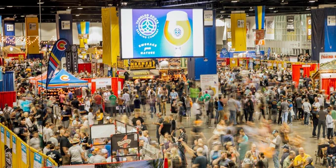 An aerial image of the Great American Beer Festival, with crowds of people walking among festival booths and large screens featuring photos of beer.