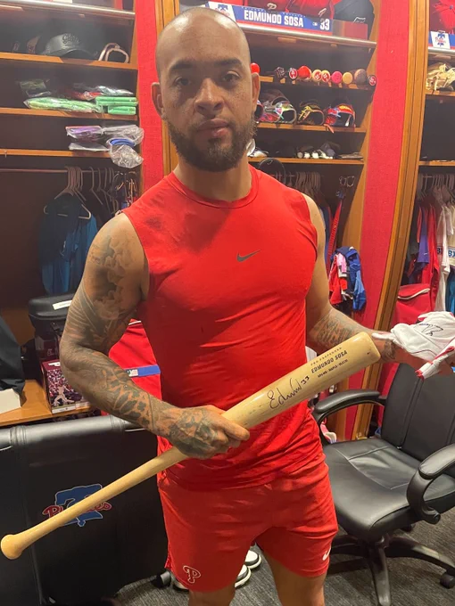 Edmundo Sosa poses for a picture in the Phillies clubhouse with an autographed bat and a pair of batting gloves. He's wearing a red sleeveless shirt with matching shorts.