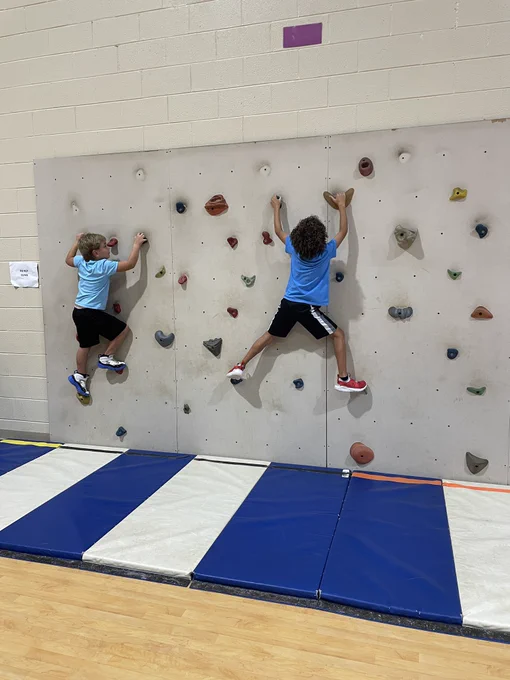 Two boys grasp climbing holds as they move across a climbing wall with a safety mat below the wall.