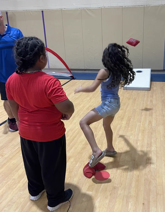 A girl throws a corn hole bag toward the corn hole board while another child looks on.