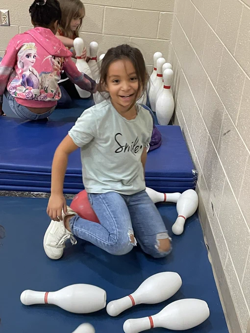 A girl sits on a ball and smiles while she sets back up the bowling pins in a make-shift bowling lane. Two other children set up pins in the background.