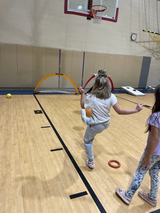 A girl kicks a ball toward a goal set up near the wall of the gym. Another child stands behind her waiting for her turn.