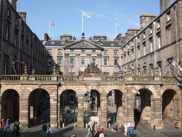 Colour image of the City Chambers in Edinburgh. It is a clear day with a blue sky. Walking by the building, on the Royal Mile, are a number of people.