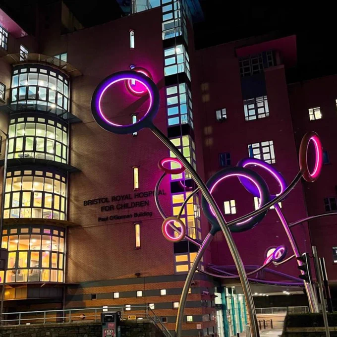 The front of Bristol Royal Hospital for Children at night. The hoops on the sculpture in front of the entrance are lit up in pink.