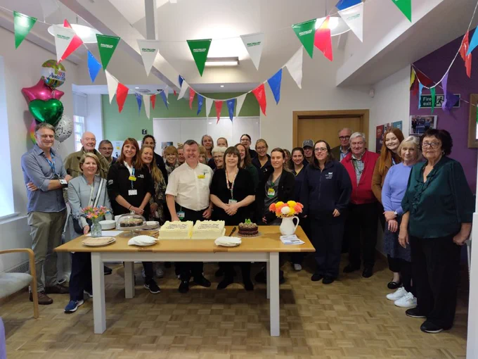 A group of people gather together, stood behind a table, where there are two large cakes. There are balloons behind them and bunting above. 