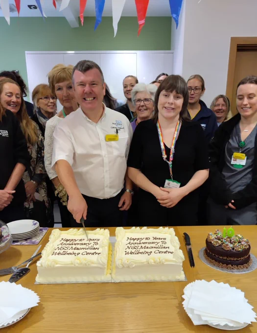 Two people stand in front of three cakes, one holding a knife to cut it and smiling at the camera. Behind them a group of people stand, smiling. On the two large cakes is the message "Happy 10 years anniversary to NGS Macmillan Wellbeing Centre"
