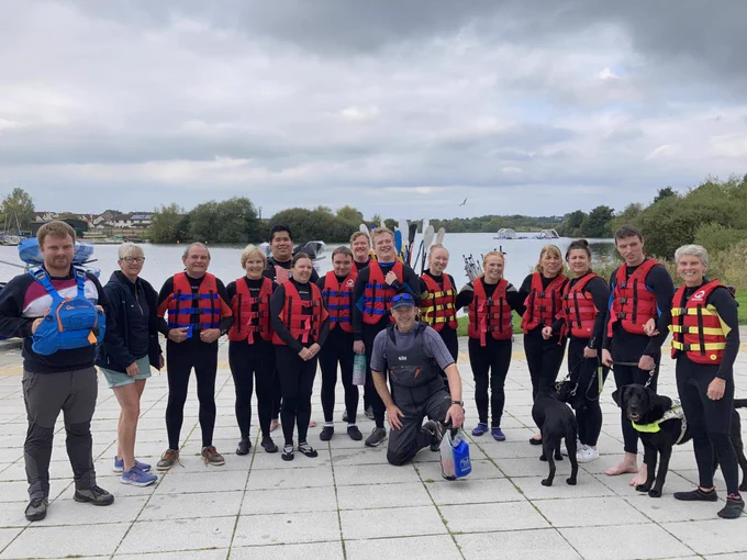 Image shows a group of  17 people a lot of whom are dressed in wet suits, standing in front of the lake and smiling. Theire are also two guide dogs Nemo and Wilma in the photo.
