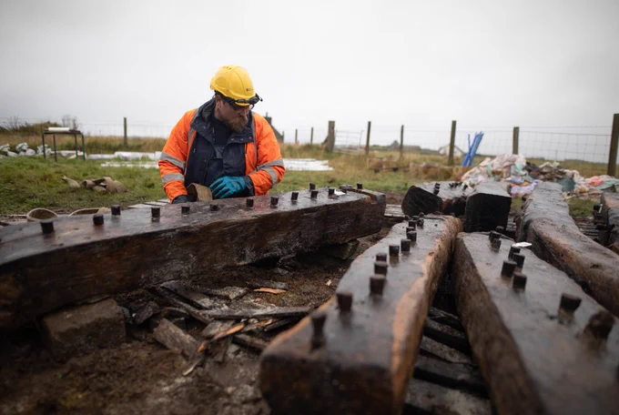 Ben crouches next to the timbers and fastens a belt around them. This will help hoist them into the tank.
