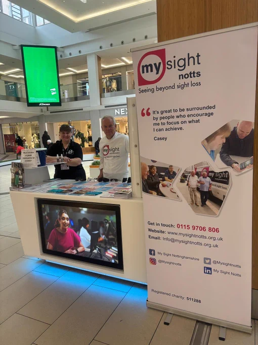 Photo shows: Sarah (Sight loss advisor) and David Cartwright (Trustee) from My Sight Notts, standing behind a branded display and banner with lots of information and leaflets to showcase what we do.