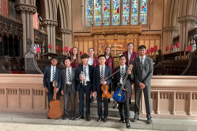 A diverse group of smiling Bolton School students in uniform: six boys on the front row, four of them holding musical instruments, and four girls on the back row. The instruments are a classical guitar, a cornet, a violin and a blue electric guitar. They are standing in front of and on the steps leading to the chancel of a church, with carved stone pillars and choir seating visible behind them to each side,  and in the background the altar and impressive stained glass window.