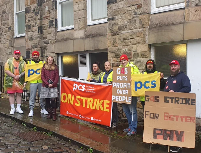 9 men and women standing on a wet pavement with PCS flags, banner and home-made placards.
