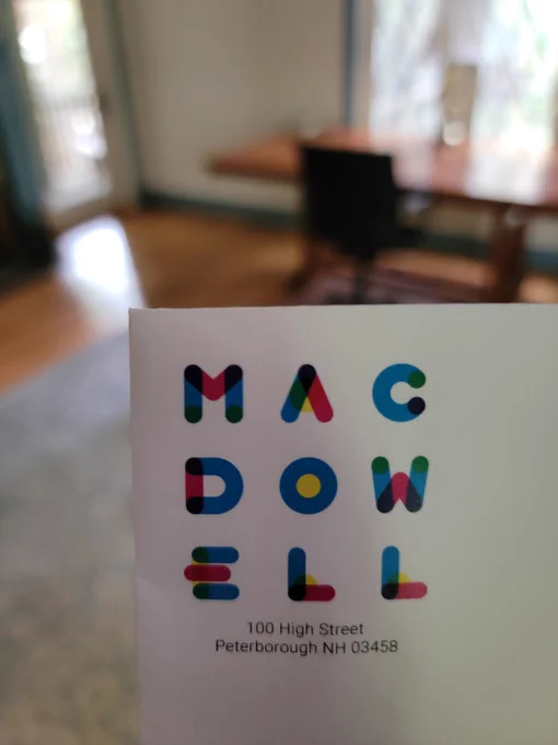 An envelope with the colorful logo of MacDowell Artist Residency printed on it. Diana (not seen) holds the envelope up. Behind it, you can see a big desk that's right in front of a window