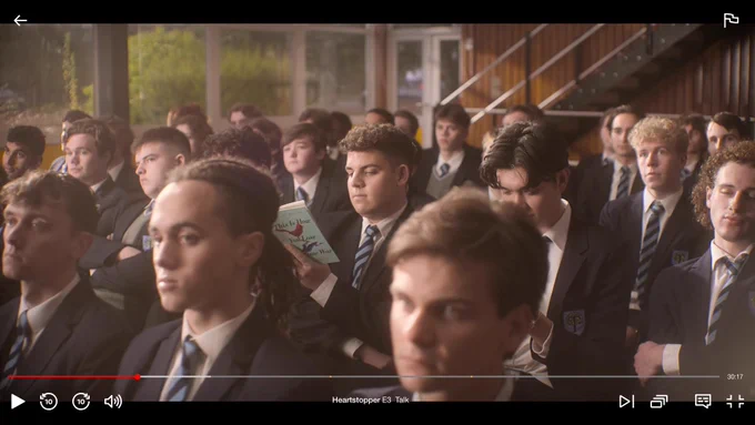 Screenshot of a crowd scene from S3 of HEARTSTOPPER in which Isaac is reading a copy of THIS IS HOW YOU LOSE THE TIME WAR, a book I wrote half of!