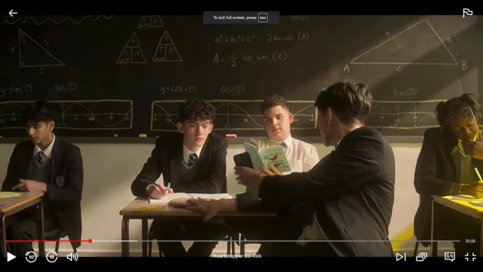 Screenshot of another scene from Heartstopper, in a classroom, in front of a blackboard, with the same boy reading the same book.