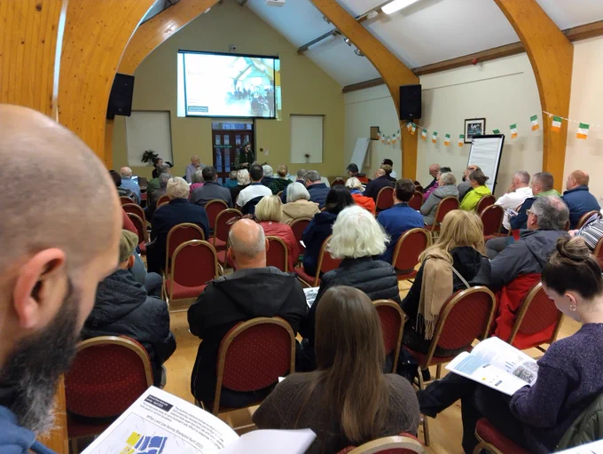 A photograph of a meeting with speakers and members of the audience reading a report booklet.