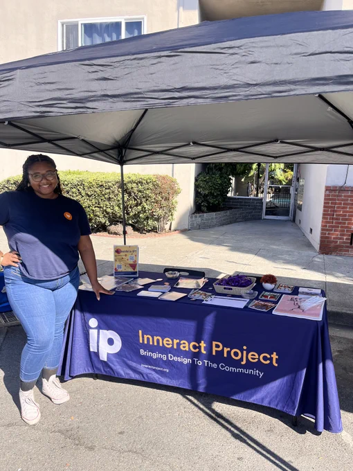 A young Black woman in a blue T-shirt and jeans poses next to a merch table under a black tent and in front of a low-rise stucco apartment building. The table is draped in a navy blue cloth and says "Inneract Project" on the front in gold.