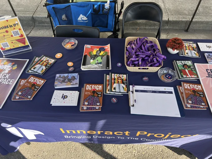 The merch table draped in navy blue cloth. On it are colorful information postcards, student-designed posters, purple branded lanyards, and a QR code to scan with the IP website (InneractProject.org).