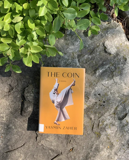 Yasmin Zaher's novel THE COIN rests on a large stone with plants encroaching the top left corner.