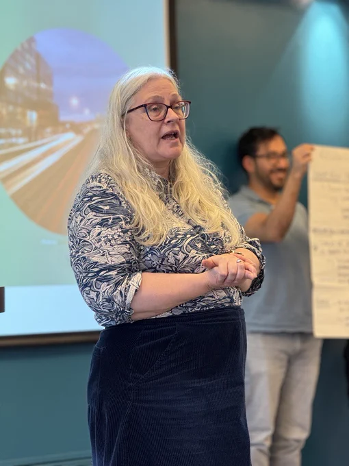 Kate Pangbourne at the EDRC Whole Centre Meeting, speaking in front of an audience. She is standing in a room with a teal wall behind her.