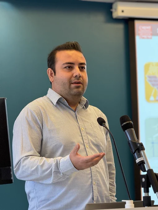 Mehmet Takci giving a presentation at the EDRC Whole Centre Meeting, speaking in front of an audience. He is standing in a room with a teal wall and a projector screen behind him.