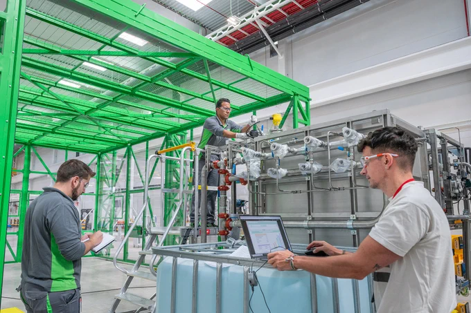 Three workers in an industrial setting with large equipment, one on a raised platform, one inspecting machinery, and one using a laptop. Bright green overhead structures add color to the scene.
