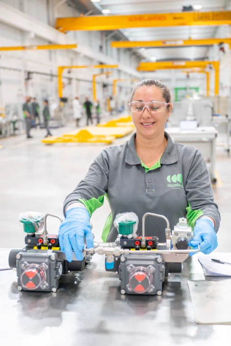 A worker at facility assembling industrial equipment, wearing safety glasses and a gray uniform, stationed at a workbench in a well-lit factory.