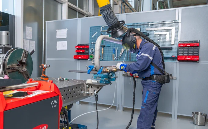 A worker wearing safety gear operates a welding machine in an industrial setting with various tools and equipment.