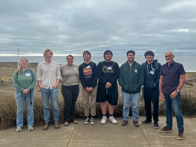 A group of five students and three professors stand in a row, looking at the camera. Behind them is a coastal view shrouded in clouds.
