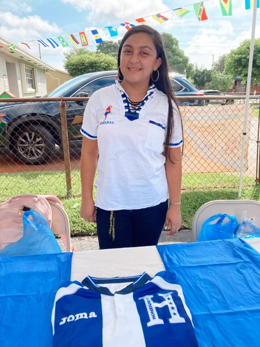 Woman standing behind a table with a blue and white Honduras soccer jersey on it. Flags are strung overhead.