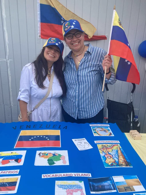 Two people wearing Venezuelan flag baseball caps stand in front of a table with Venezuelan-themed items and a large Venezuelan flag in the background.

