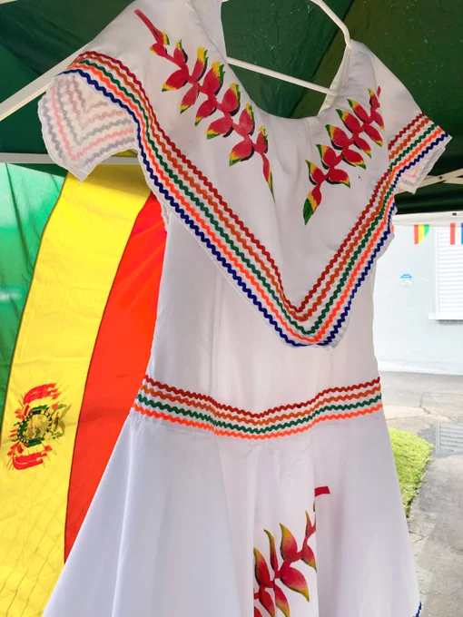 White dress with colorful embroidery and painted red flowers, displayed under a green canopy with the Bolivian flag in the background.