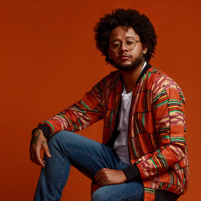 Emicida sitting on a stool wearing a colorful striped jacket and glasses, with a neutral expression, against an orange background.