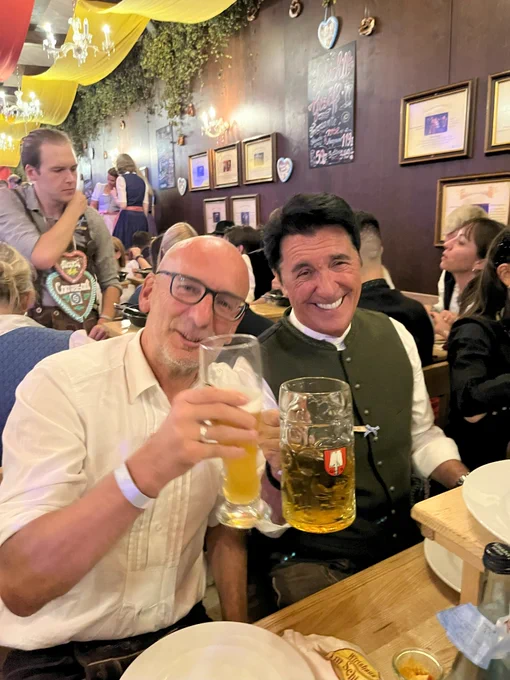 Two individuals enjoying beers at a busy festival, seated at a table with large beer mugs, one wearing a traditional vest and the other in a casual shirt.