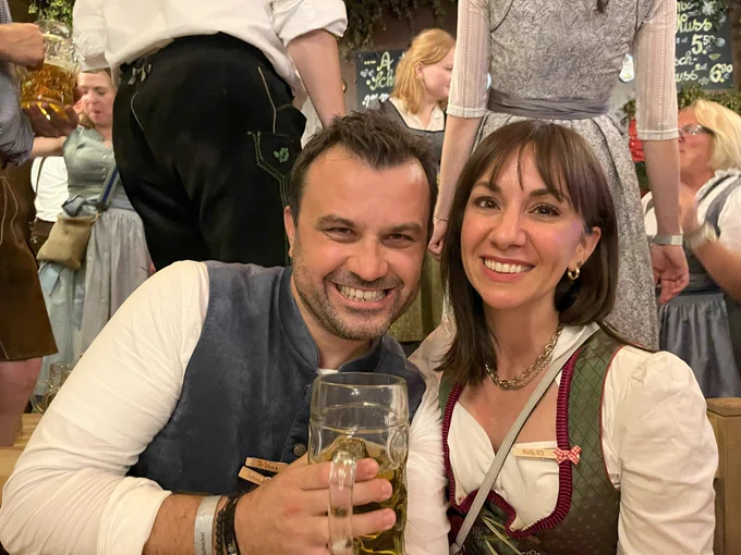 Two people smiling, dressed in traditional Bavarian outfits and holding a beer mug, at a festive event.