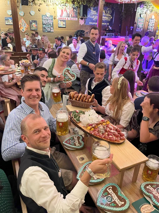 Group of people enjoying food and drinks at Oktoberfest, seated at a crowded beer hall with traditional decorations.