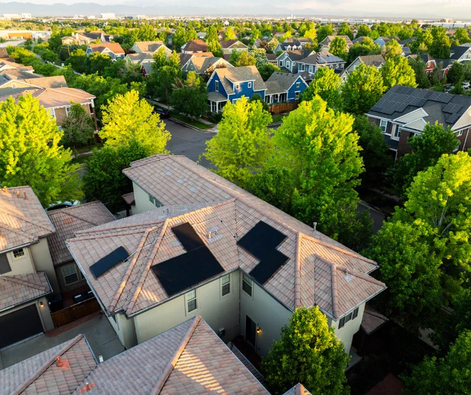 Aerial view of a suburban neighborhood with closely spaced houses, surrounded by lush green trees under a clear sky. The main house has solar panels on it.